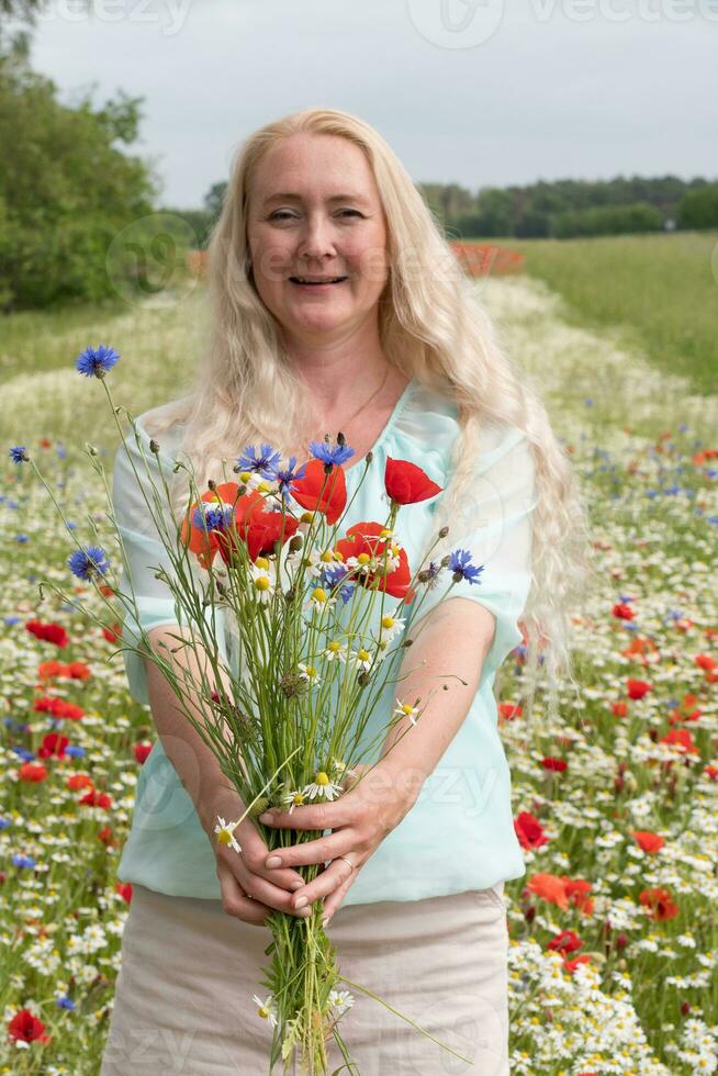 lindo meia idade Loiras mulher carrinhos entre uma floração campo do papoilas foto