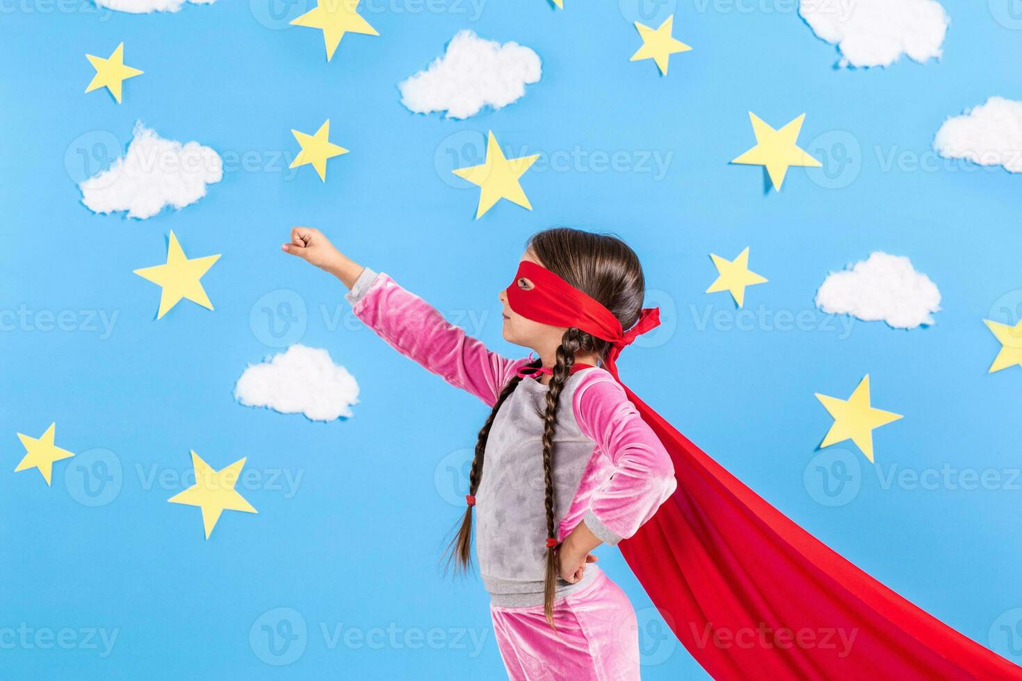 pequeno criança tocam Super heroi. criança em a fundo do brilhante azul parede com branco nuvens e estrelas . menina poder conceito. foto