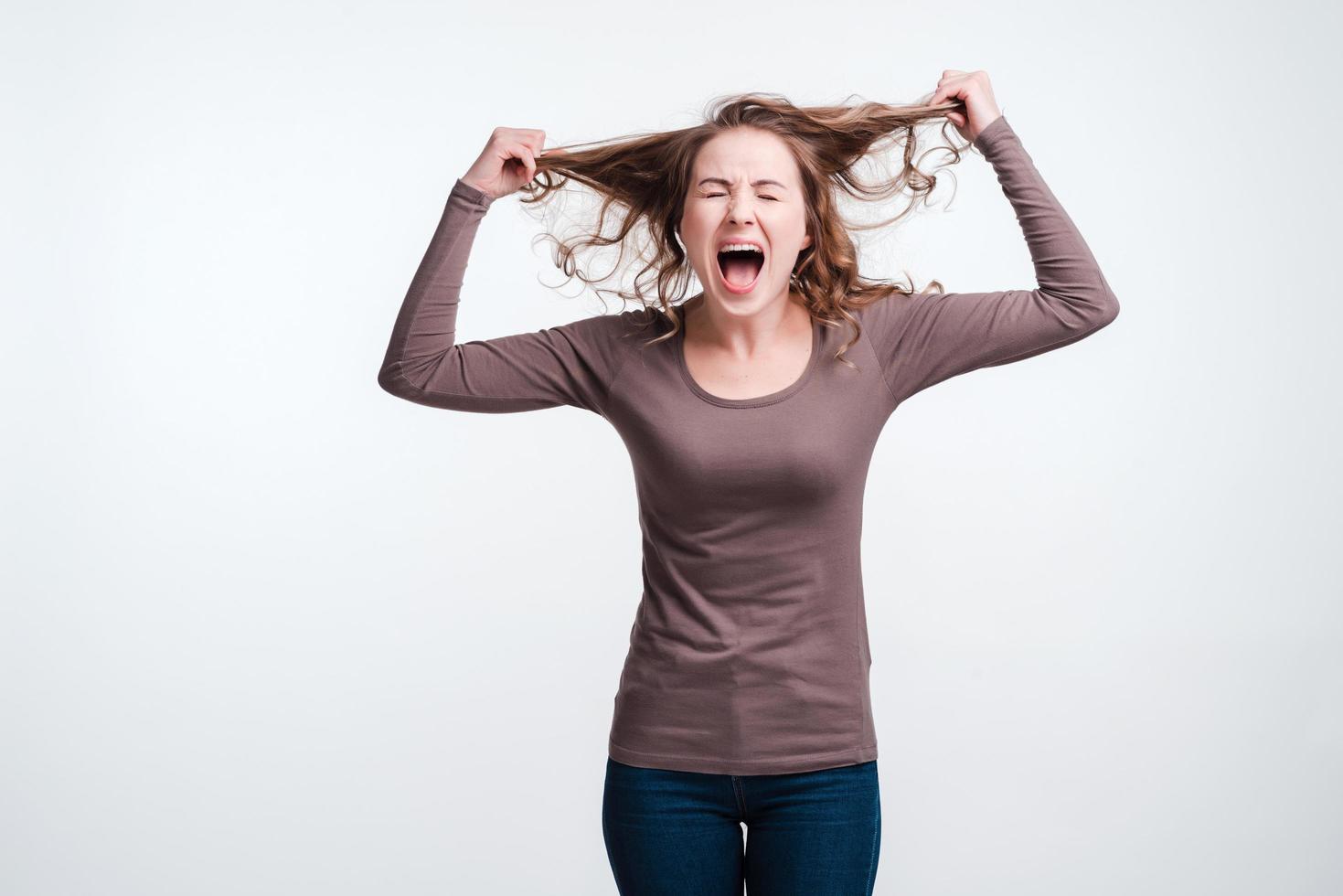 Mulher jovem gritando animada em pé sobre um fundo branco e segurando o cabelo foto