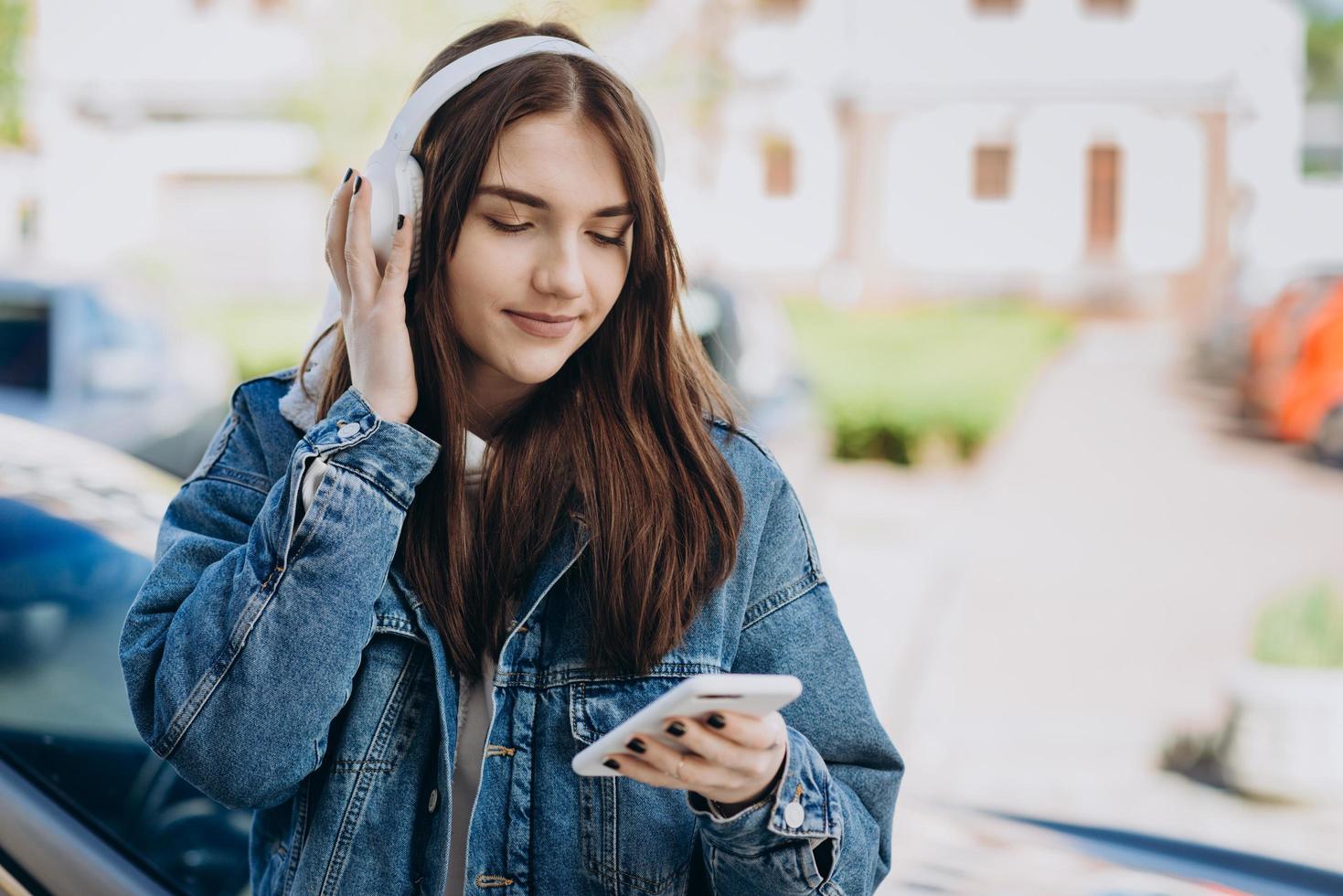 linda jovem morena de branco, fones de ouvido sem fio e com um telefone nas mãos, em um plano de fundo da cidade foto