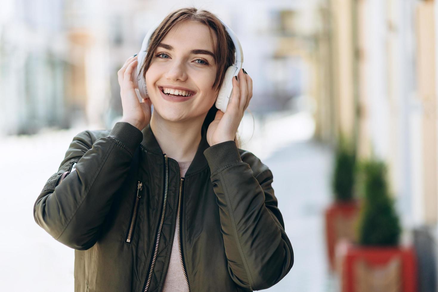 adorável senhora em pé na cidade e ouvindo a música favorita em fones de ouvido. retrato ao ar livre da sonhadora menina europeia usa fones de ouvido brancos. foto