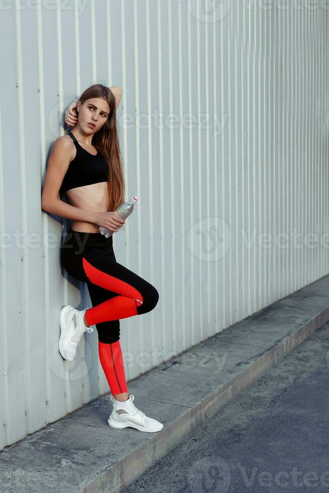 fêmea atleta bebendo água enquanto em pé de uma cinzento muro. foto
