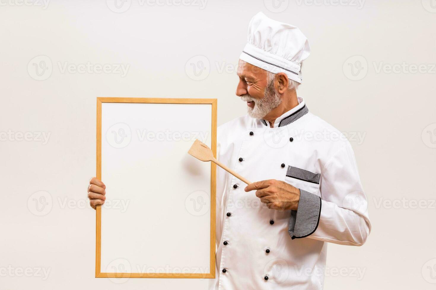 retrato do Senior chefe de cozinha mostrando com de madeira colher quadro branco em cinzento fundo. foto