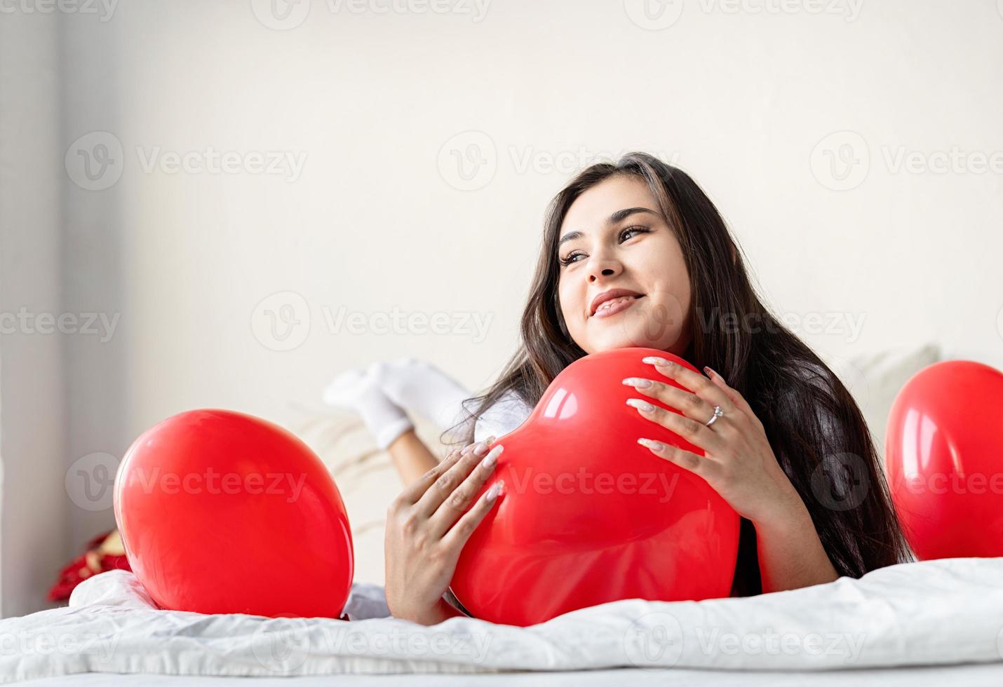 jovem mulher morena feliz deitada na cama com balões em forma de coração vermelho foto