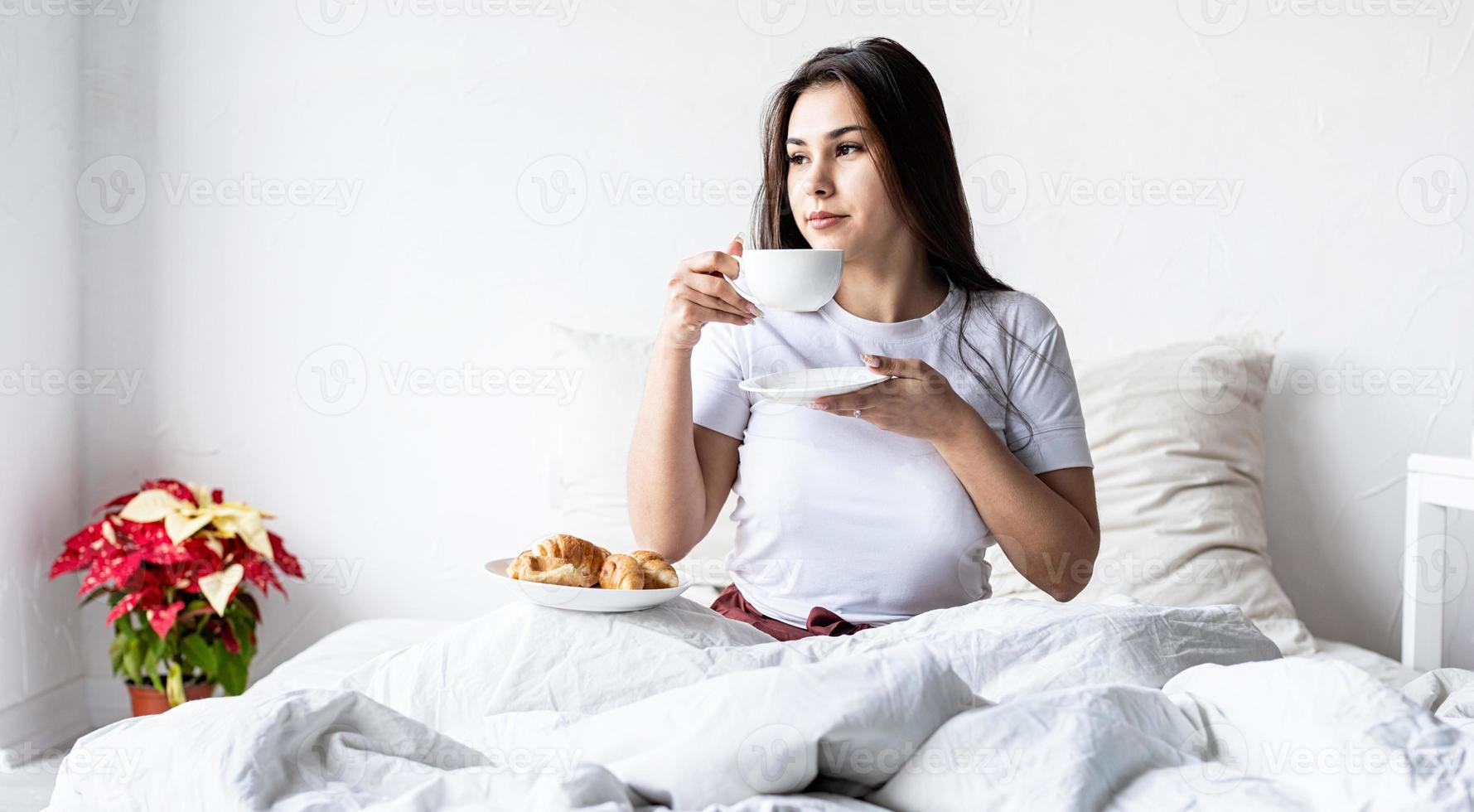 jovem morena sentada acordada na cama com balões em forma de coração vermelho e decorações, bebendo café comendo croissants foto