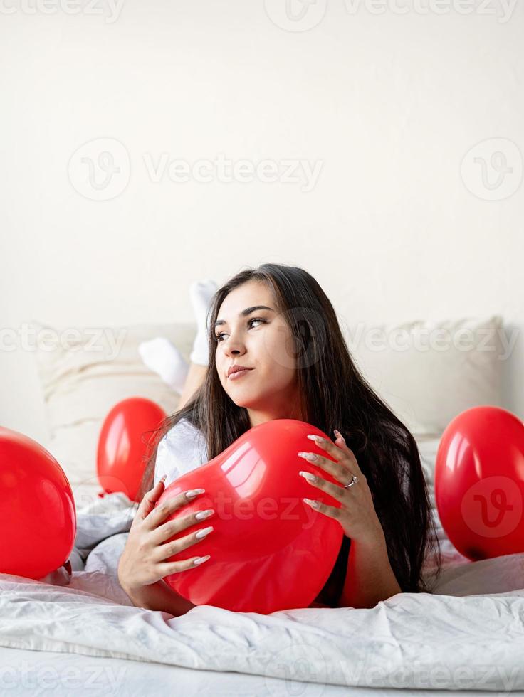 jovem mulher morena feliz deitada na cama com balões em forma de coração vermelho foto