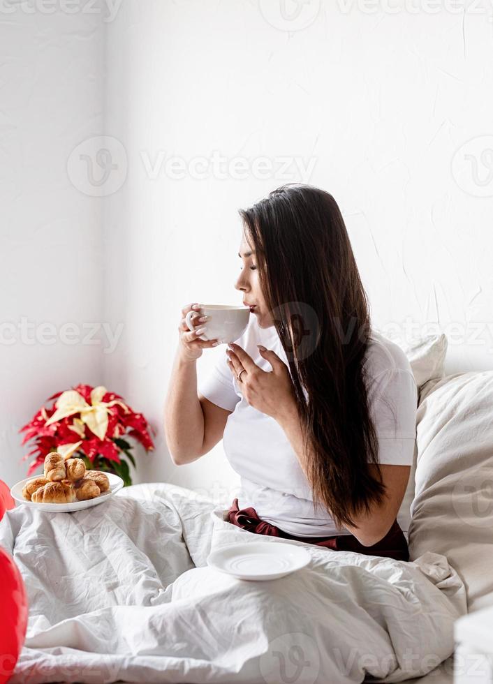 jovem morena sentada acordada na cama com balões em forma de coração vermelho e decorações, bebendo café comendo croissants foto