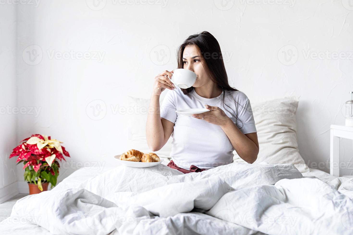 jovem morena sentada acordada na cama com balões em forma de coração vermelho e decorações, bebendo café comendo croissants foto