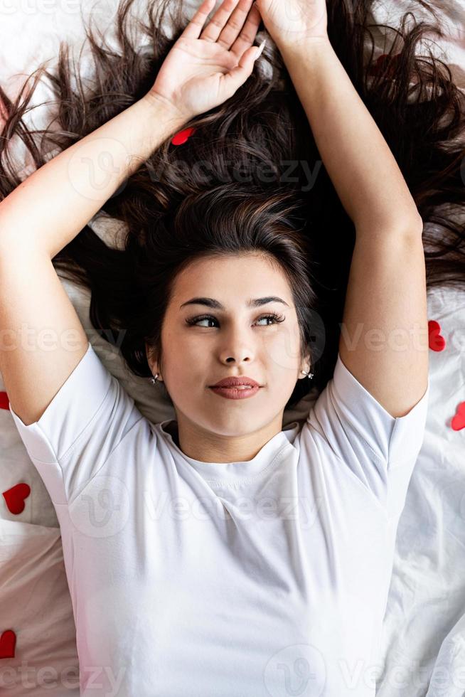 jovem mulher morena feliz deitada na cama com enfeites em forma de coração vermelho foto