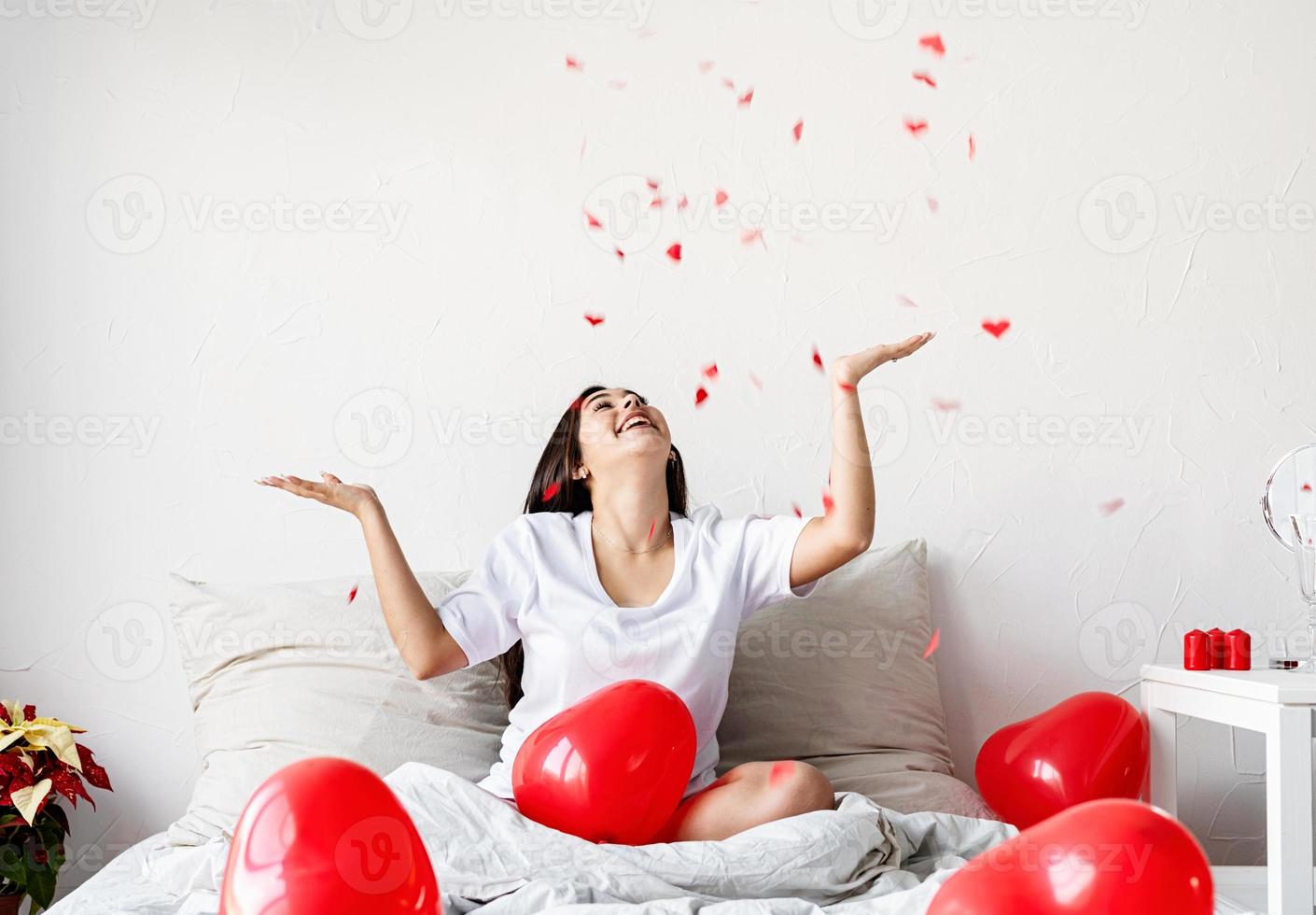 jovem morena feliz sentada na cama com balões em forma de coração vermelho jogando confete para o alto foto