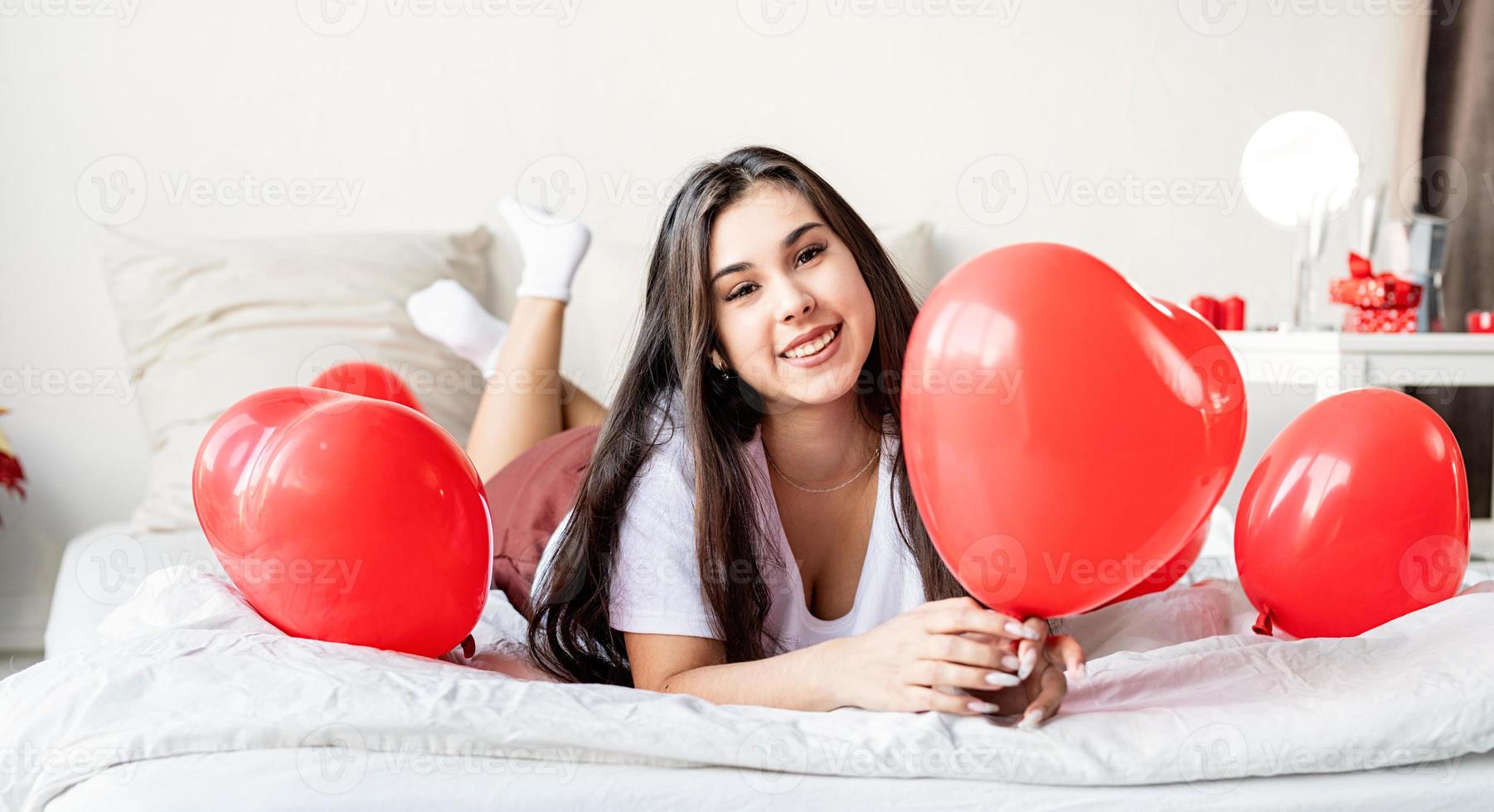 jovem mulher morena feliz deitada na cama com balões em forma de coração vermelho foto