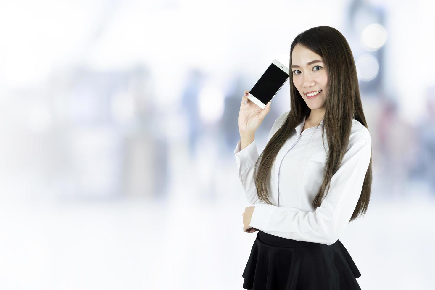 jovem linda mulher de negócios asiáticos sorrindo, segurando um telefone inteligente de tela em branco no fundo desfocado foto