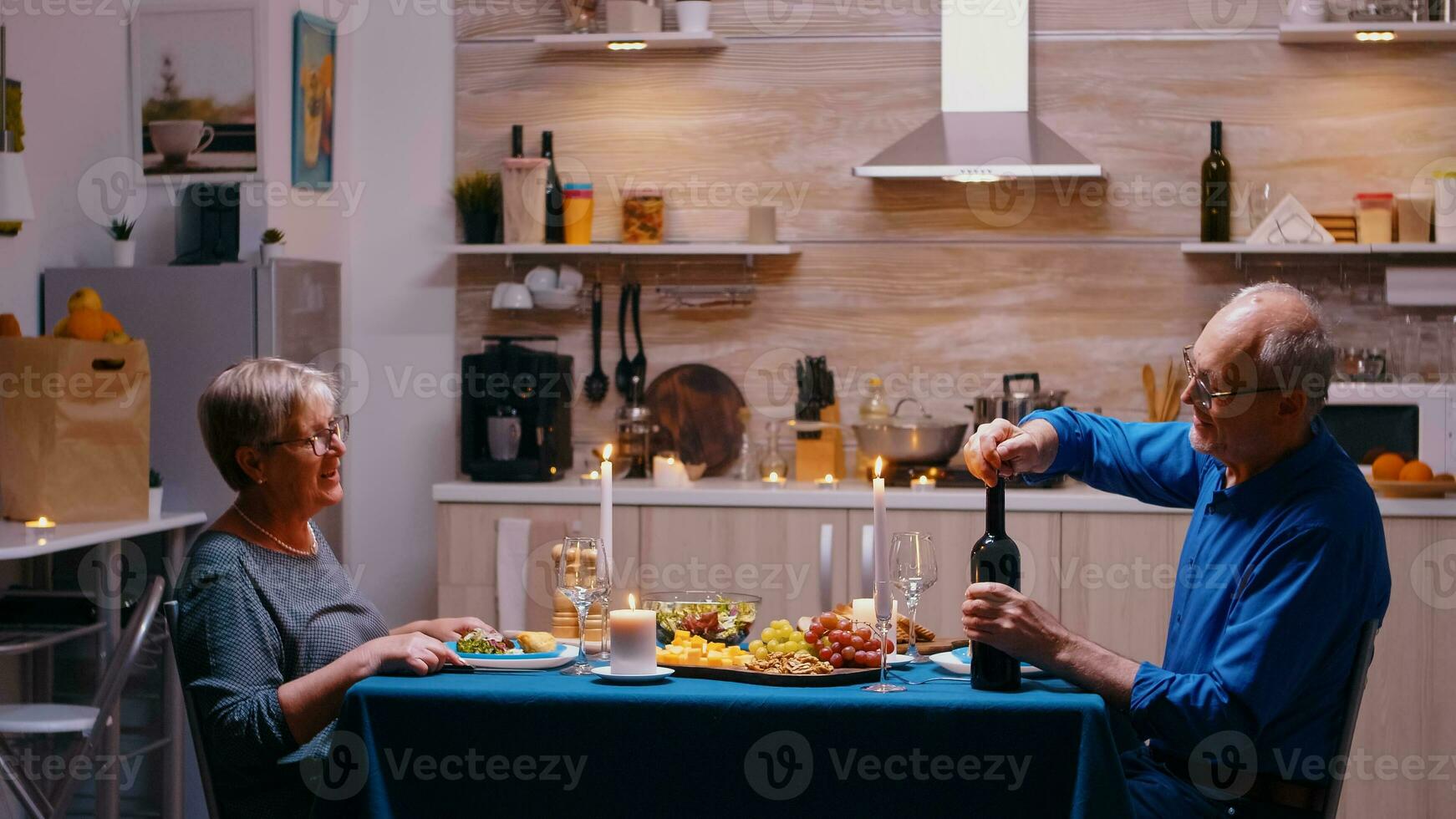 idosos homem abertura uma garrafa do vermelho vinho durante romântico jantar. Senior velho casal conversando, sentado às a mesa dentro cozinha, desfrutando a refeição, a comemorar seus aniversário às casa com saudável Comida. foto