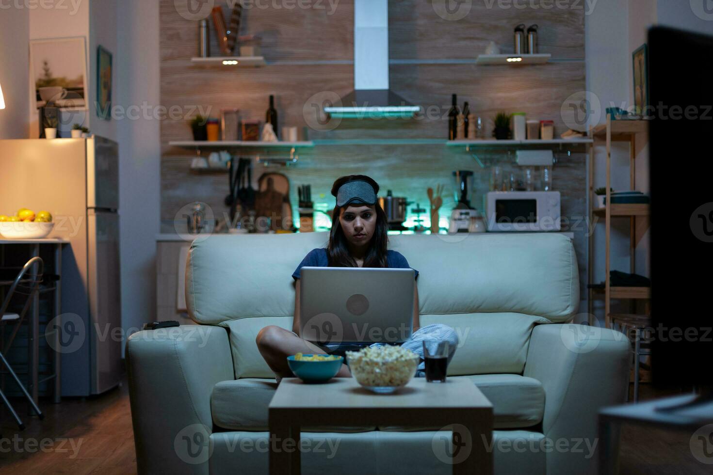 mulher com olho mascarar usando computador portátil às noite enquanto assistindo televisão e comendo lanches. feliz pessoa dentro pijamas sentado em sofá lendo escrevendo procurando navegando em caderno usando Internet verificação mails foto