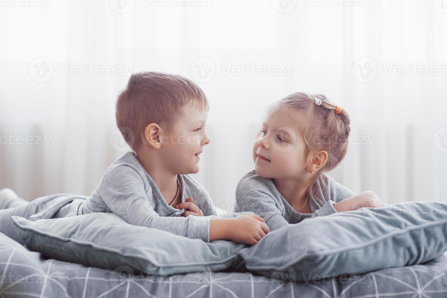 crianças brincando na cama dos pais. as crianças acordam em um quarto branco e ensolarado. menino e menina brincam de pijama combinando. roupa de dormir e roupa de cama para criança e bebê. interior do berçário para criança criança. manhã familiar foto