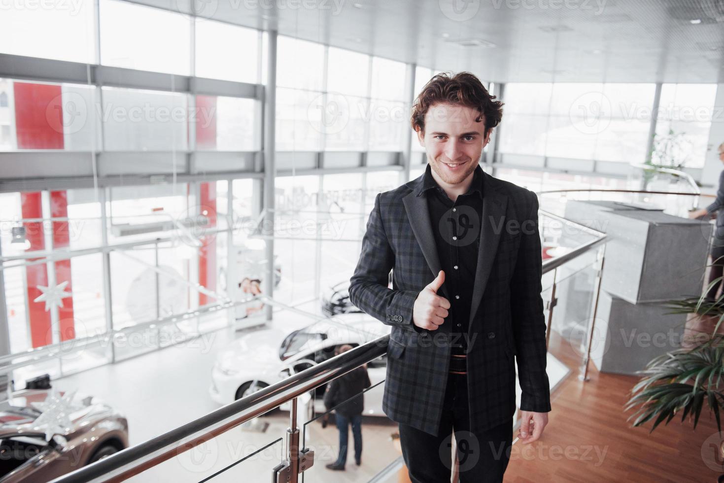 visitando concessionária de automóveis. jovem no fundo do salão do carro. homem feliz aparece o dedo. conceito de venda e aluguel de carros foto