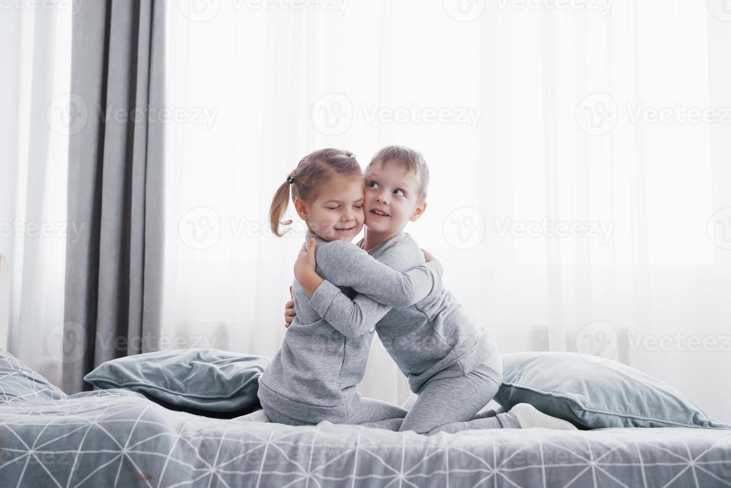 crianças felizes brincando no quarto branco. menino e menina, irmão e irmã brincam na cama de pijama. interior do berçário para crianças. pijamas e roupas de cama para bebês e crianças pequenas. familia em casa foto