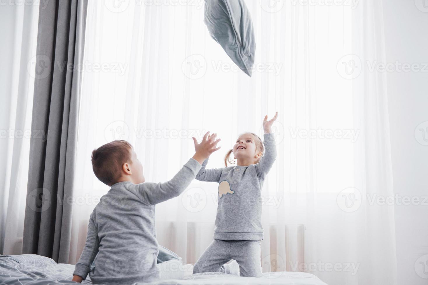 crianças brincando na cama dos pais. as crianças acordam em um quarto branco e ensolarado. menino e menina brincam de pijama combinando. roupa de dormir e roupa de cama para criança e bebê. interior do berçário para criança criança. manhã familiar foto