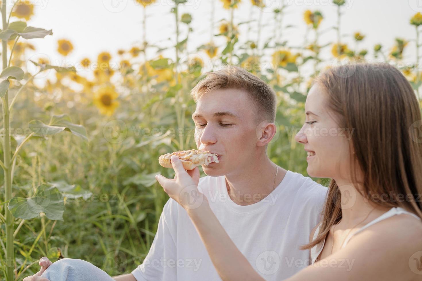 jovem casal fazendo piquenique no campo de girassol ao pôr do sol, mulher alimentando boyfrind com pizza foto