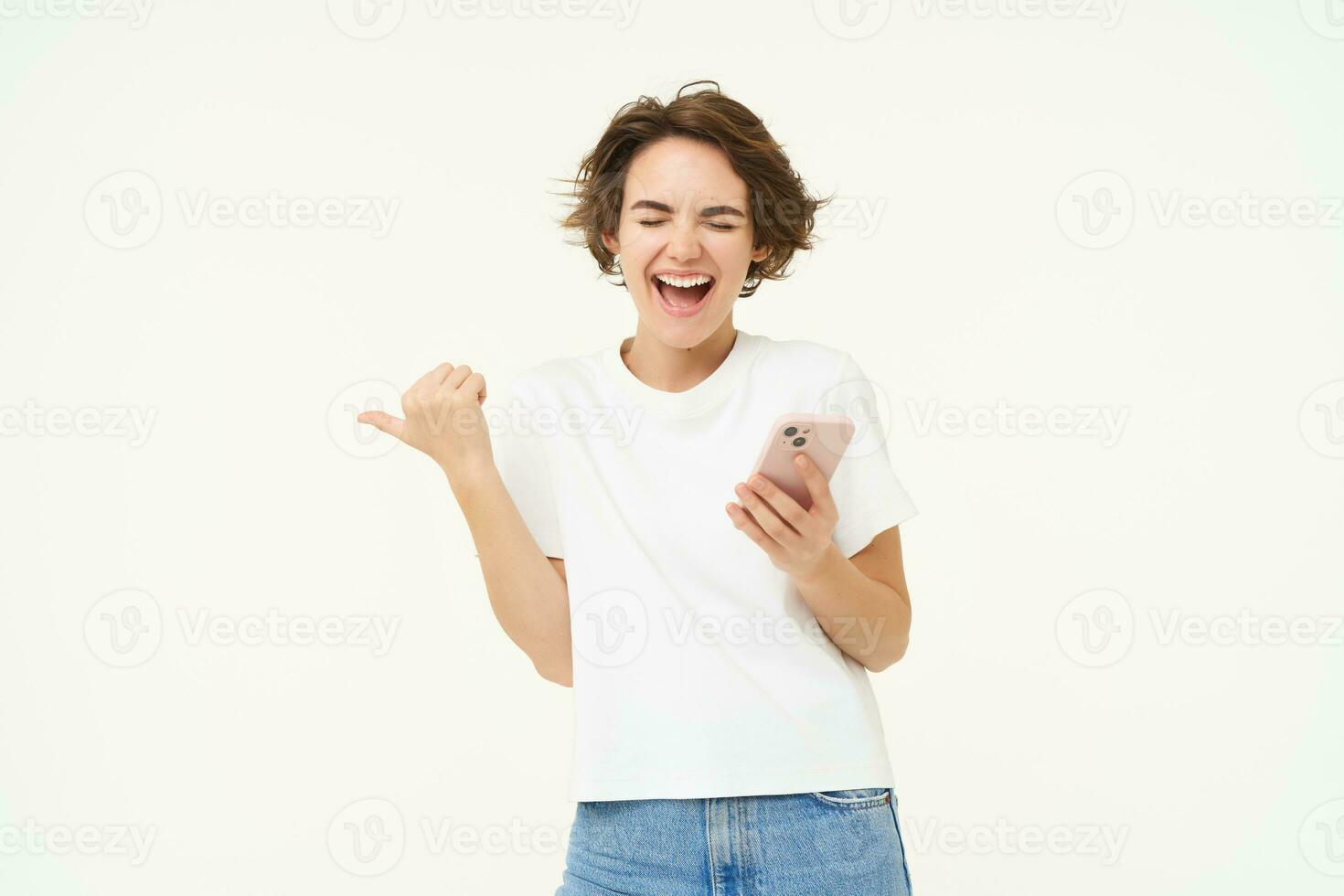retrato do lindo, moderno menina com Smartphone, apontando dedo esquerda, segurando Smartphone e rindo, posando sobre branco estúdio fundo foto