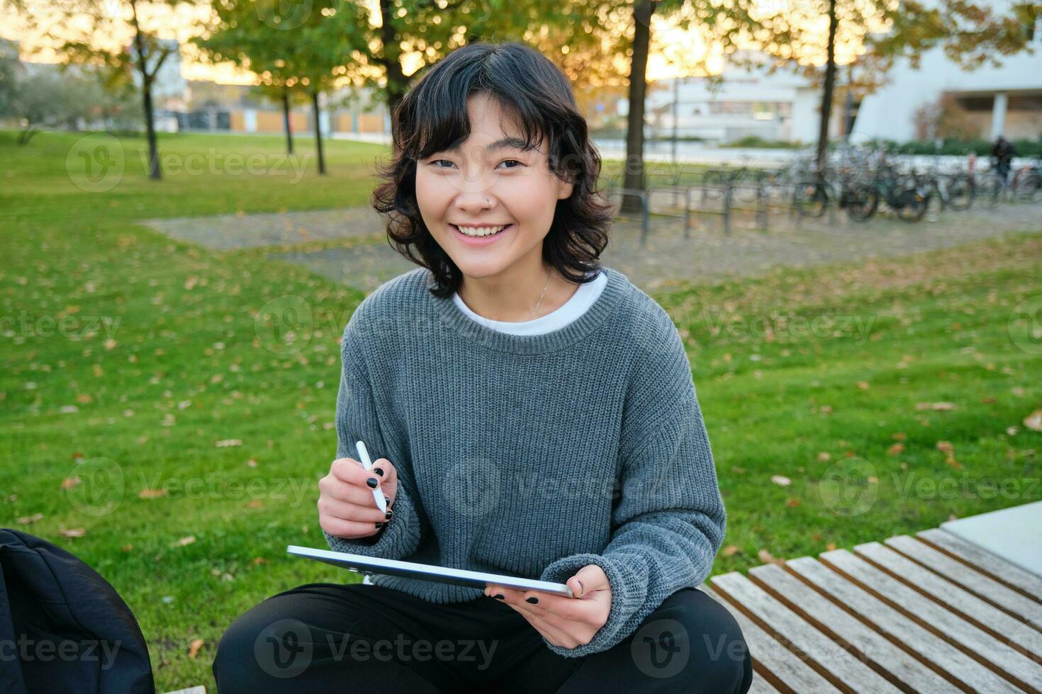 retrato do jovem sorridente coreano garota, gráfico desenhista, artista desenhando em digital tábua com uma caneta ferramenta, sentado dentro parque em fresco ar e pegador, levando notas foto