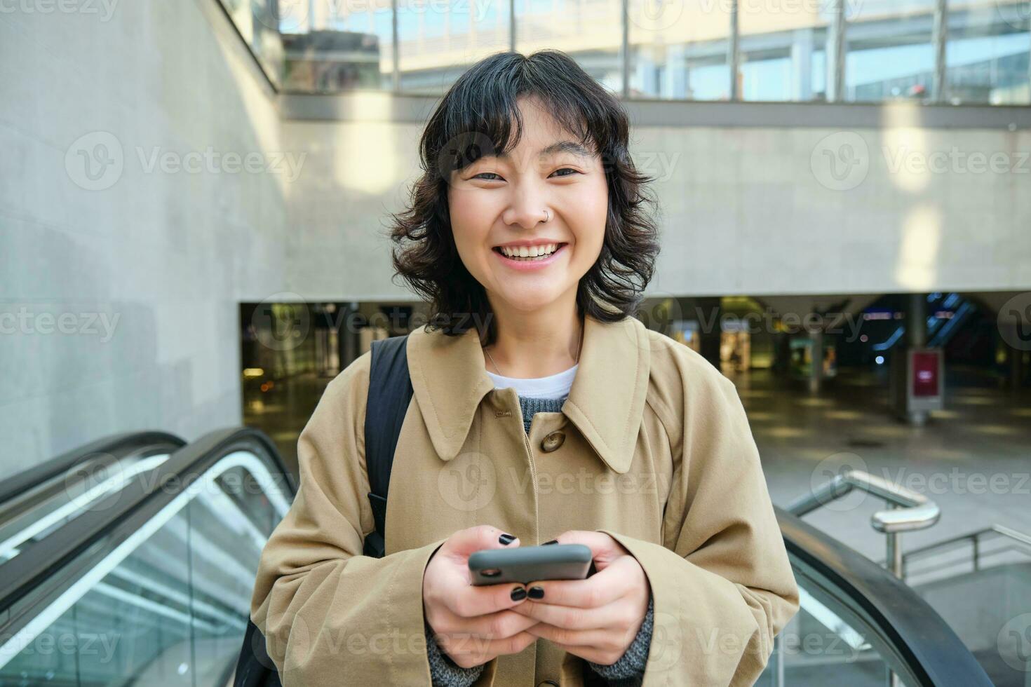 pessoas dentro cidade. jovem coreano mulher viaja por aí cidade, vai acima a escada rolante, usa dela Móvel telefone e sorrisos foto