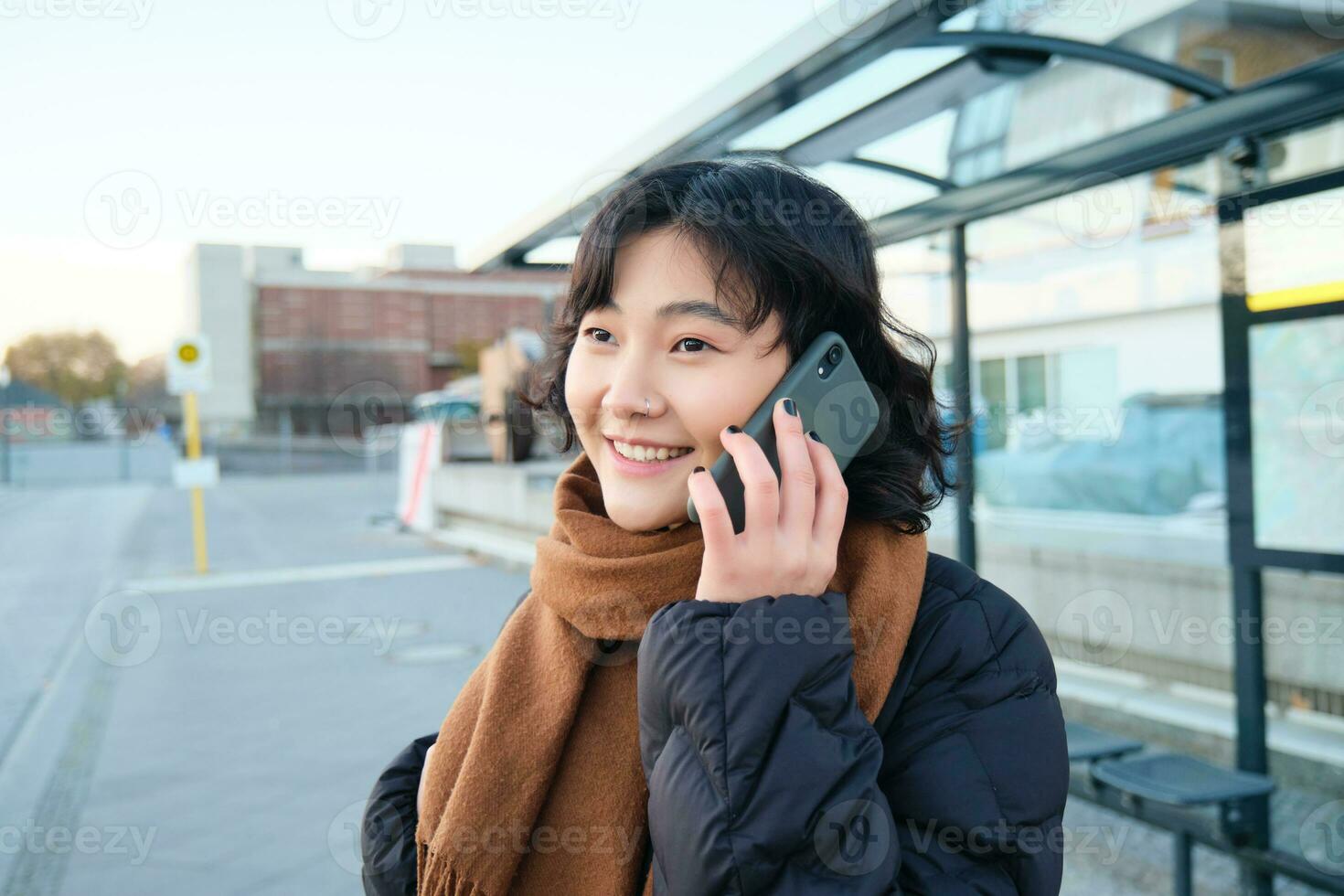 sorrindo coreano menina falando em Móvel telefone, em pé em ônibus parar, usando Smartphone, posando em estrada dentro inverno, embrulhado dentro lenço, vestindo Preto Jaqueta foto