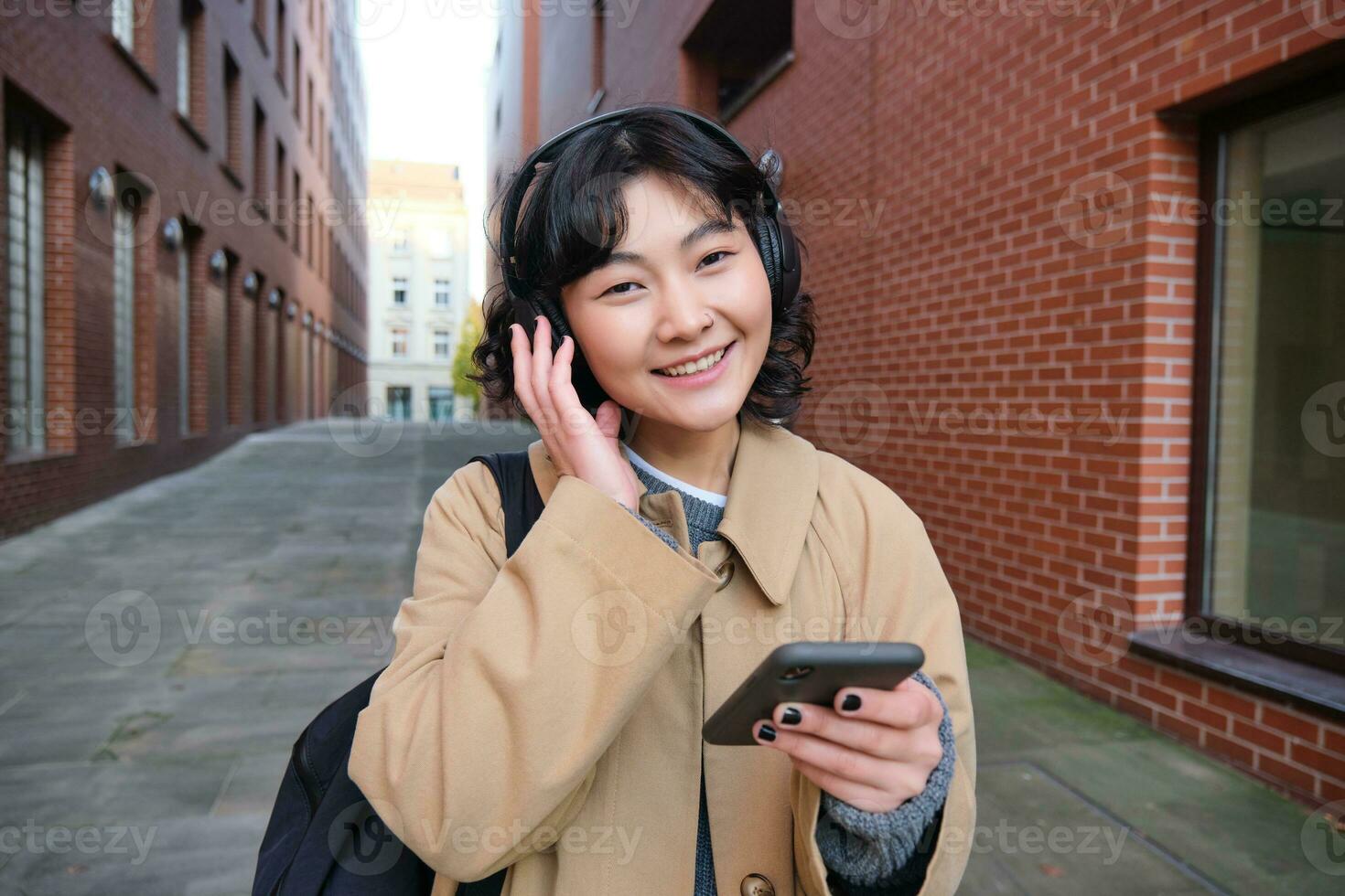 retrato do jovem coreano mulher caminhando por aí cidade com mochila e fones de ouvido, escuta música, parece às Smartphone, usa telefone inscrição em ruas foto