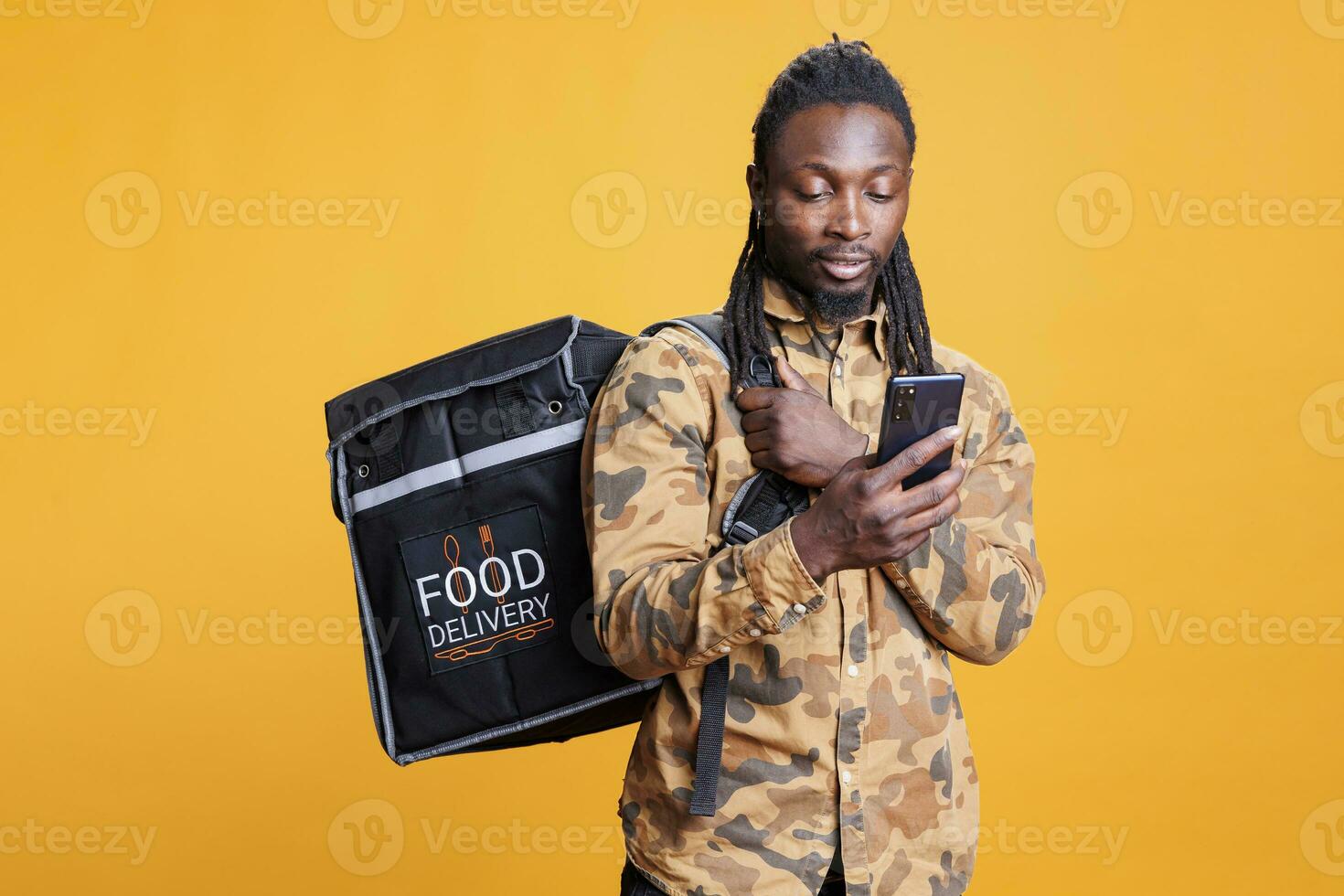 africano americano correio rastreamento cliente endereço em Smartphone antes entregando ordem durante almoço tempo. entregador segurando térmico mochila cheio com Leve embora Comida sobre amarelo fundo foto