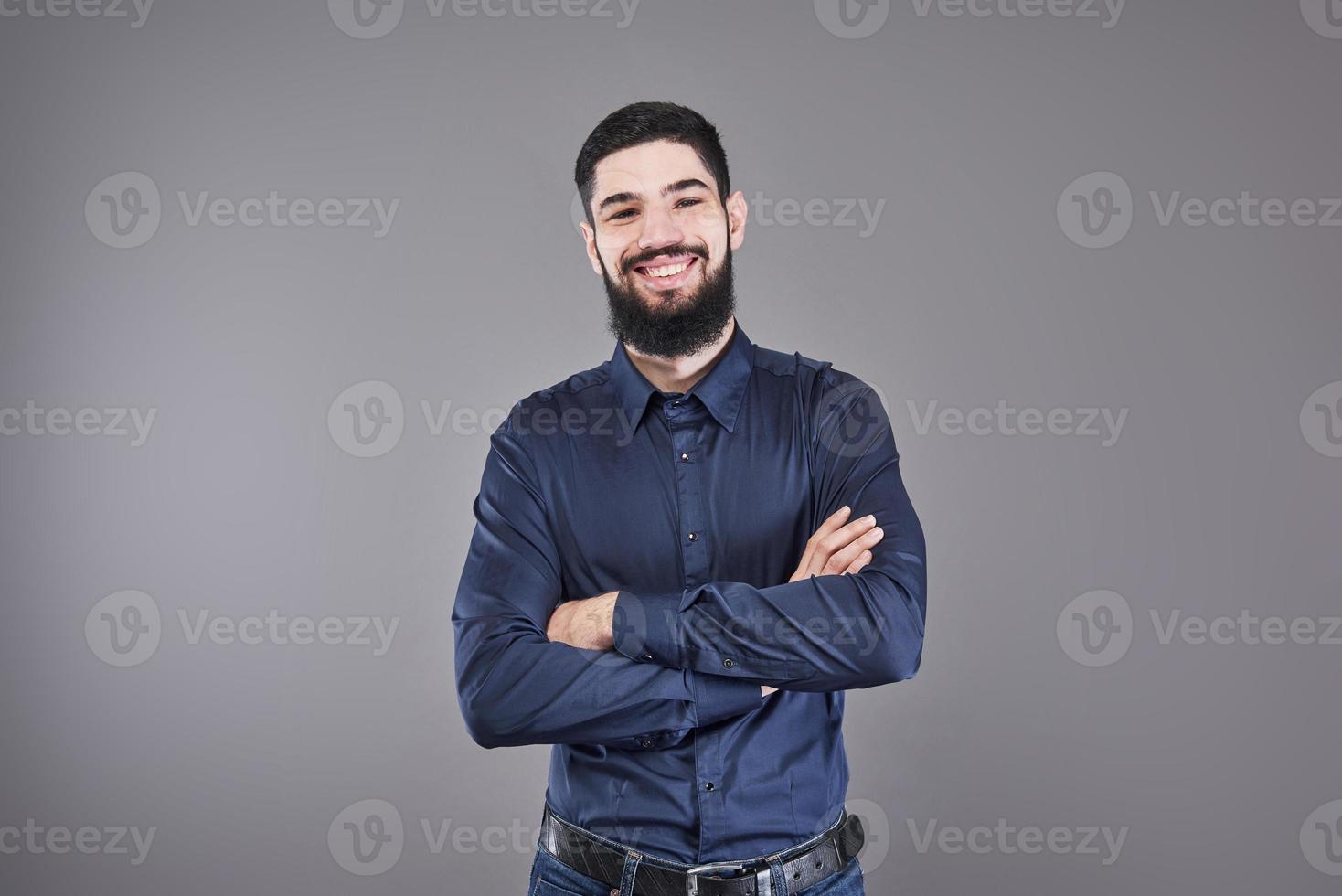 jovem bonito encostado na parede cinza com os braços cruzados. um jovem sério com barba olha para a câmera foto