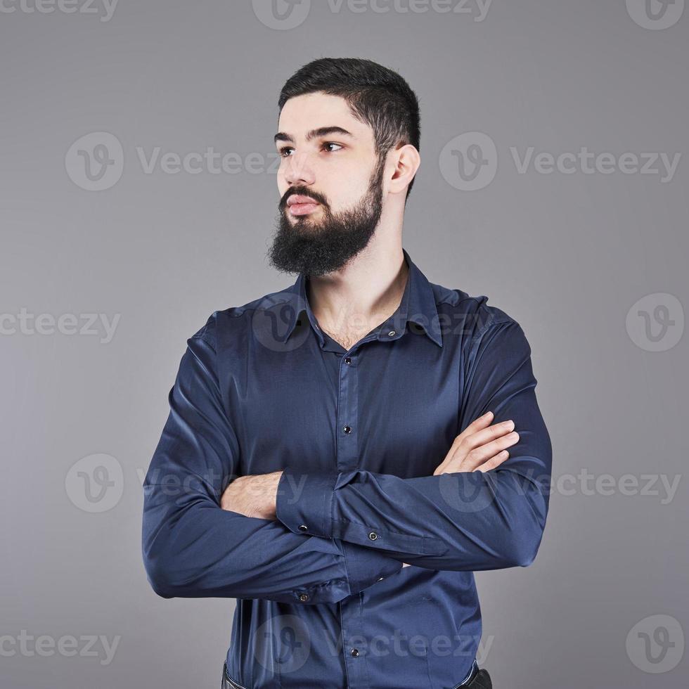 jovem bonito com barba encostado na parede cinza com os braços cruzados. foto