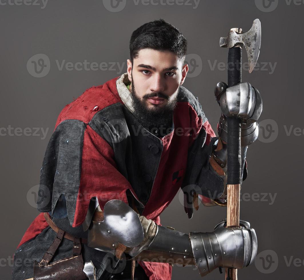 cavaleiro medieval em fundo cinza. retrato de guerreiro brutal com rosto sujo com cota de malha, roupas vermelhas e pretas e machado de batalha foto