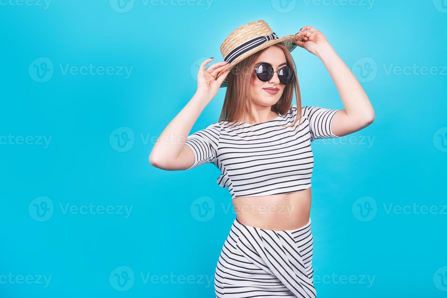 garota atraente em listras brancas e pretas, chapéu, óculos escuros, boca emocionalmente aberta sobre um fundo azul brilhante com um corpo perfeito. isolado. tiro de estúdio foto