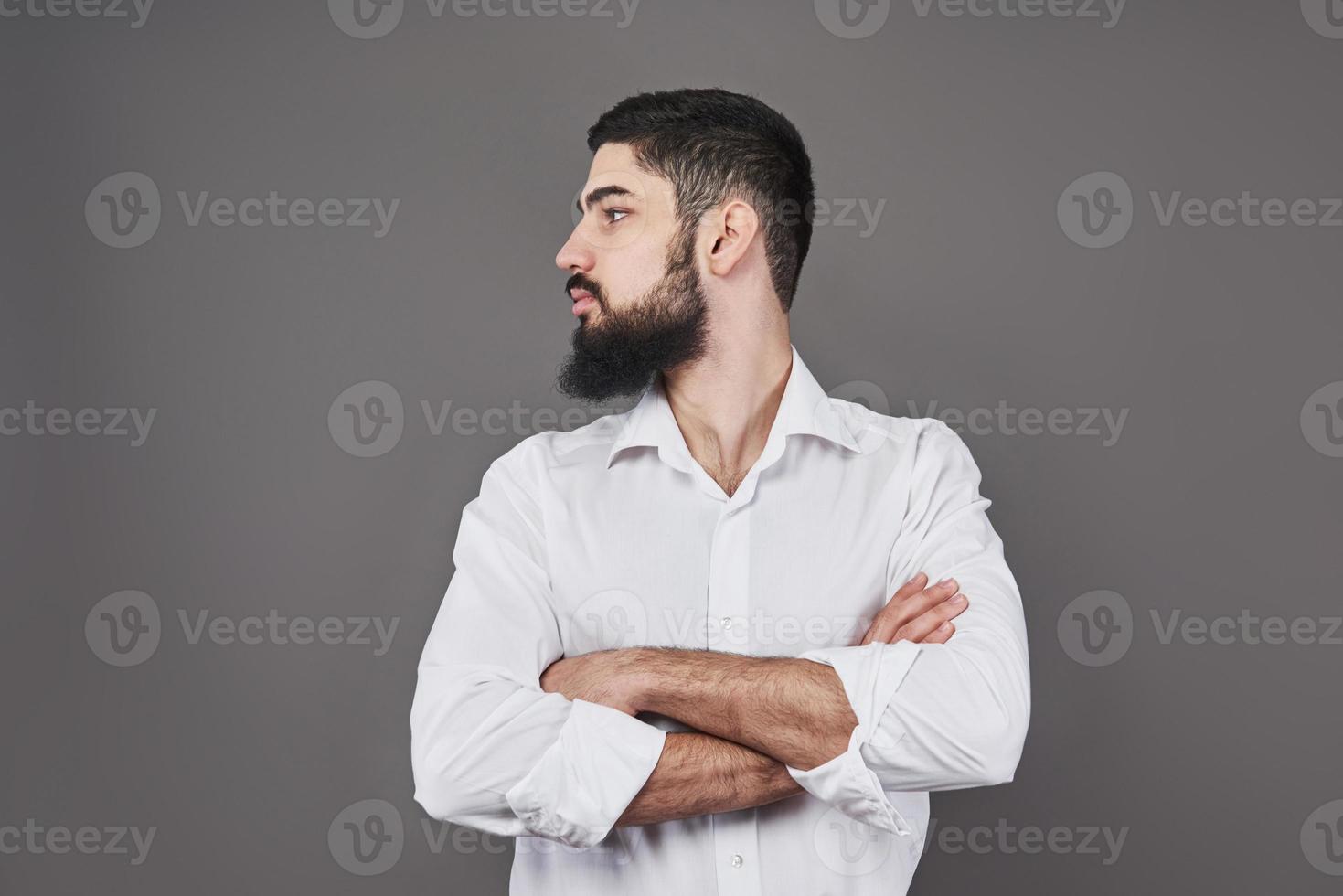 jovem bonito com barba encostado na parede cinza com os braços cruzados. foto