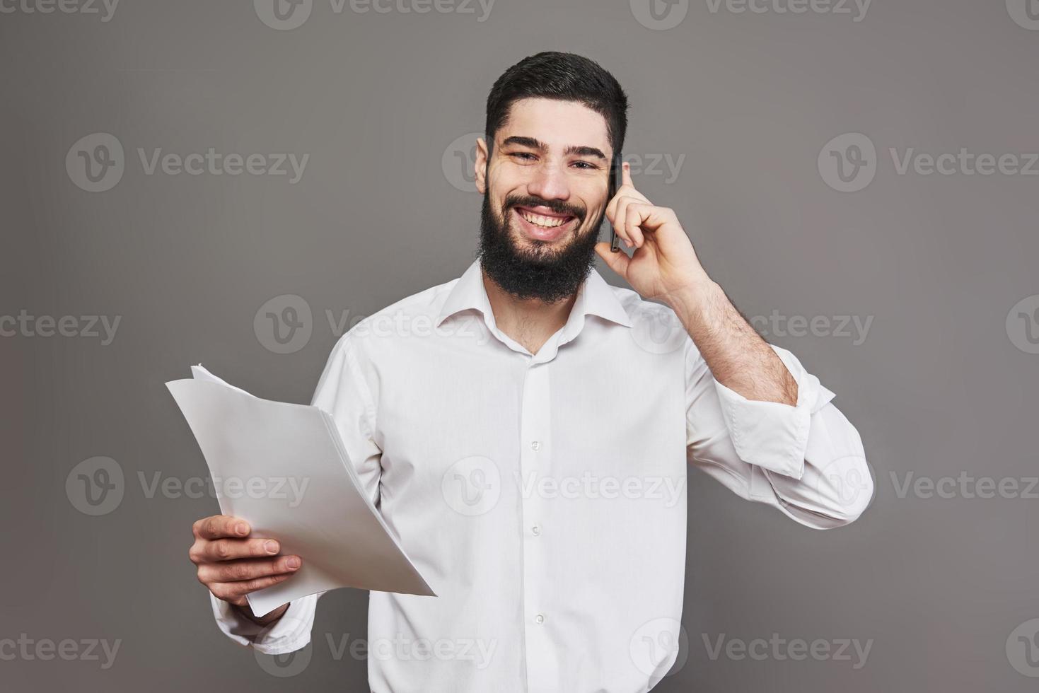 homem de negócios com barba na camisa branca segurando documentos e o telefone em fundo cinza foto