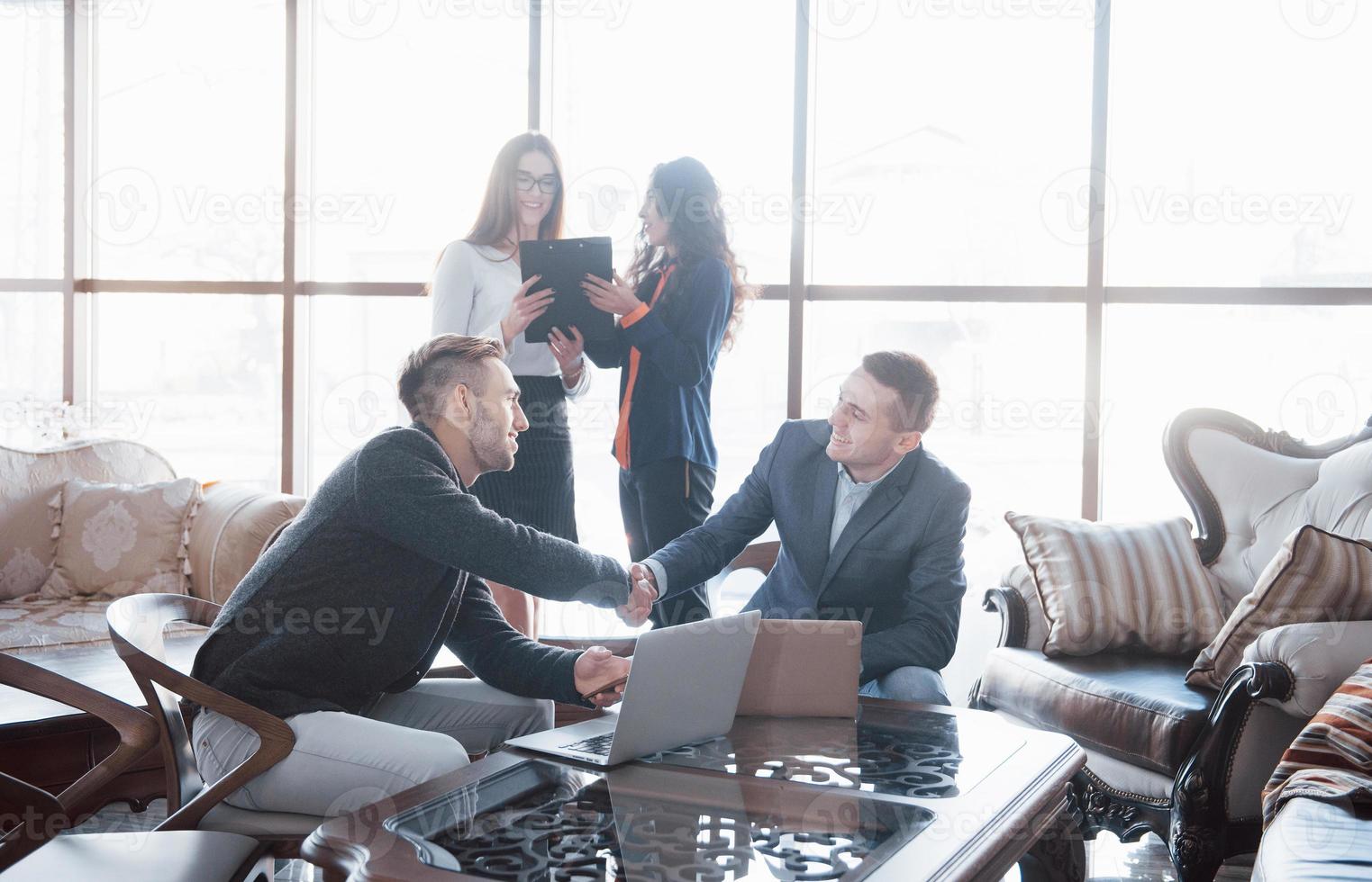 Bem-vindo a bordo de dois homens bonitos apertando as mãos com um sorriso no escritório com seus colegas de trabalho. jovem equipe de colegas de trabalho discutindo grandes negócios no moderno escritório de coworking. conceito de trabalho em equipe foto