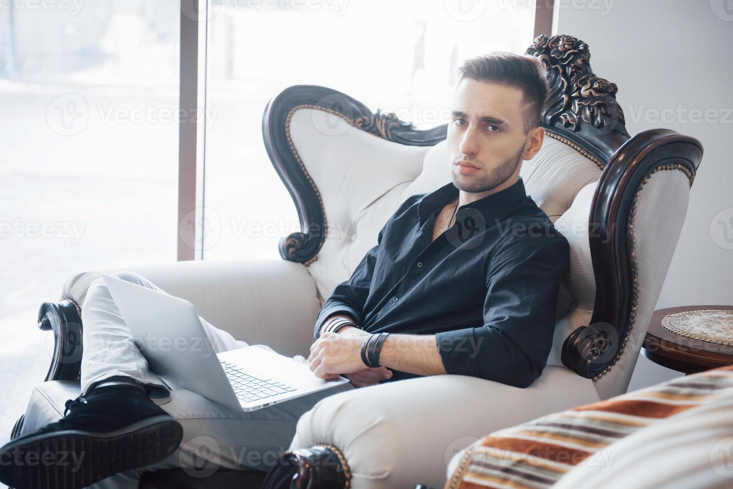 jovem empresário trabalhando em um escritório moderno. homem vestindo camisa branca e usando laptop contemporâneo. fundo de janelas panorâmicas foto