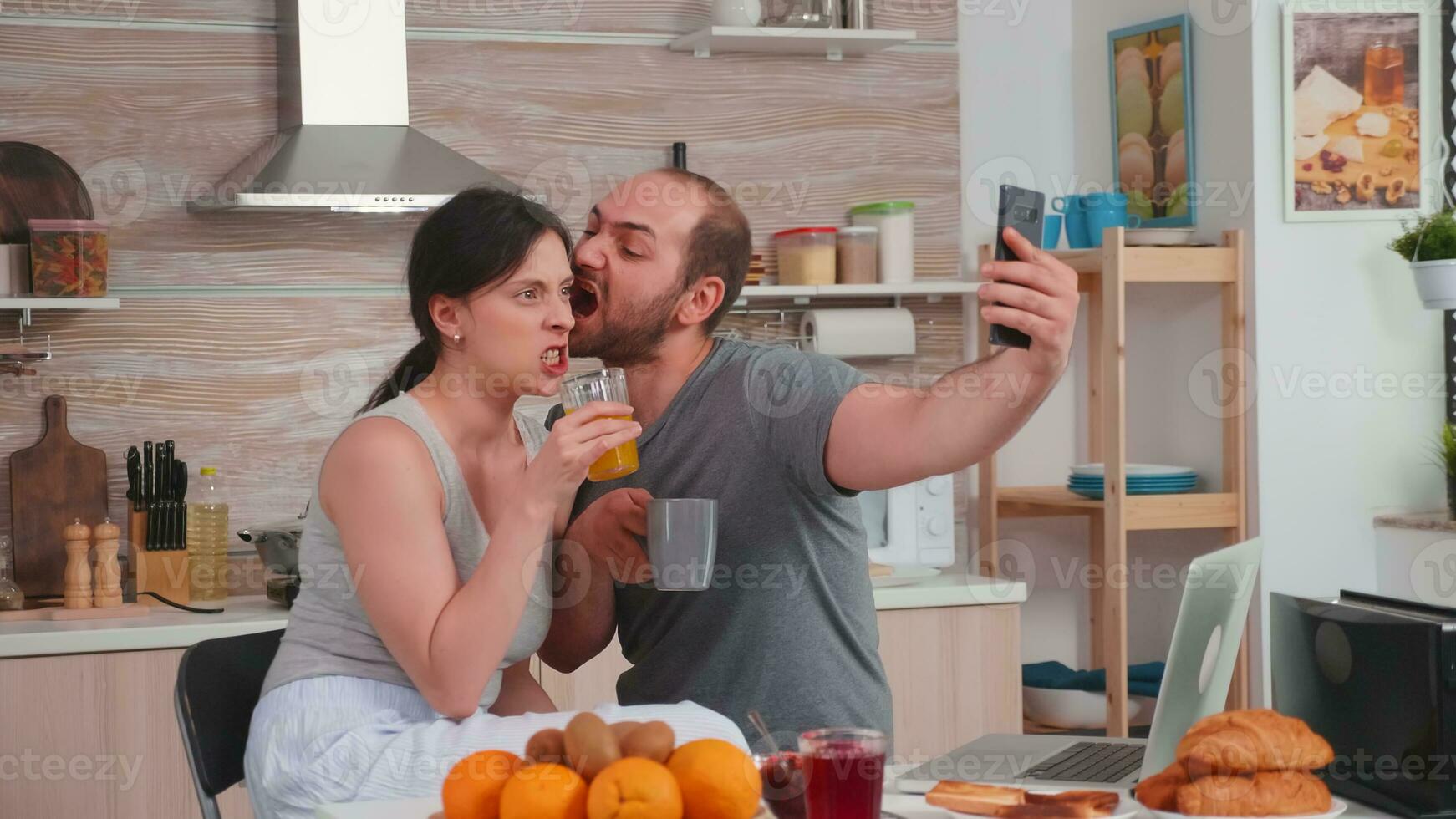 feliz jovem casal levando selfies durante café da manhã. alegre casado marido e esposa fazer engraçado rostos enquanto levando uma foto durante café da manhã dentro cozinha.