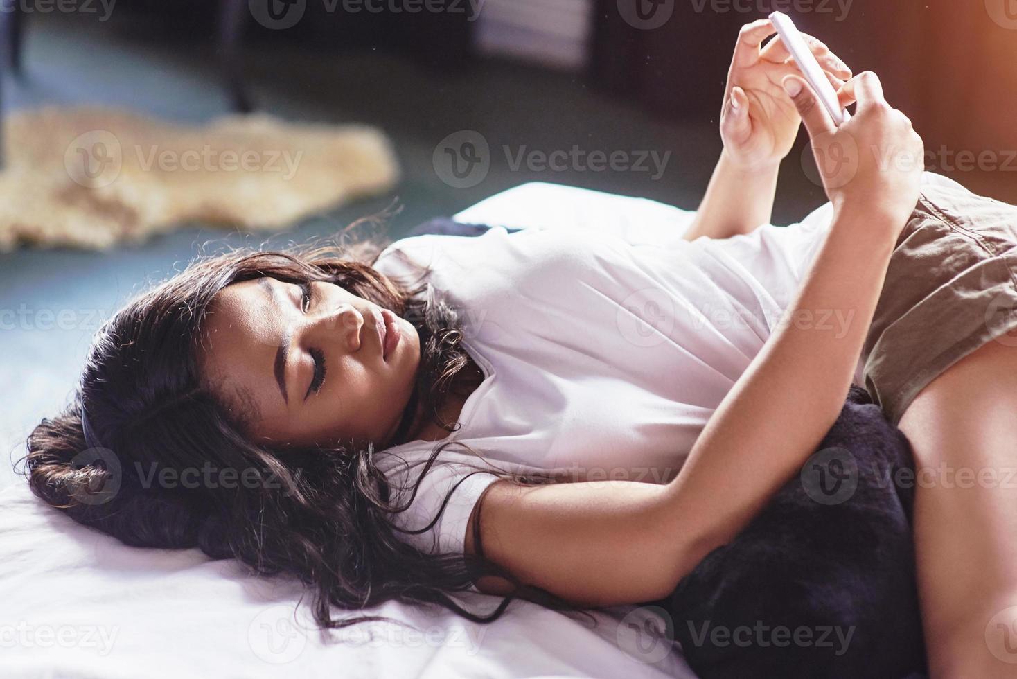 retrato de mulher bonita acordando na cama dela e olha para o telefone. verificar redes sociais, enviar sms. a garota está vestindo uma camiseta foto