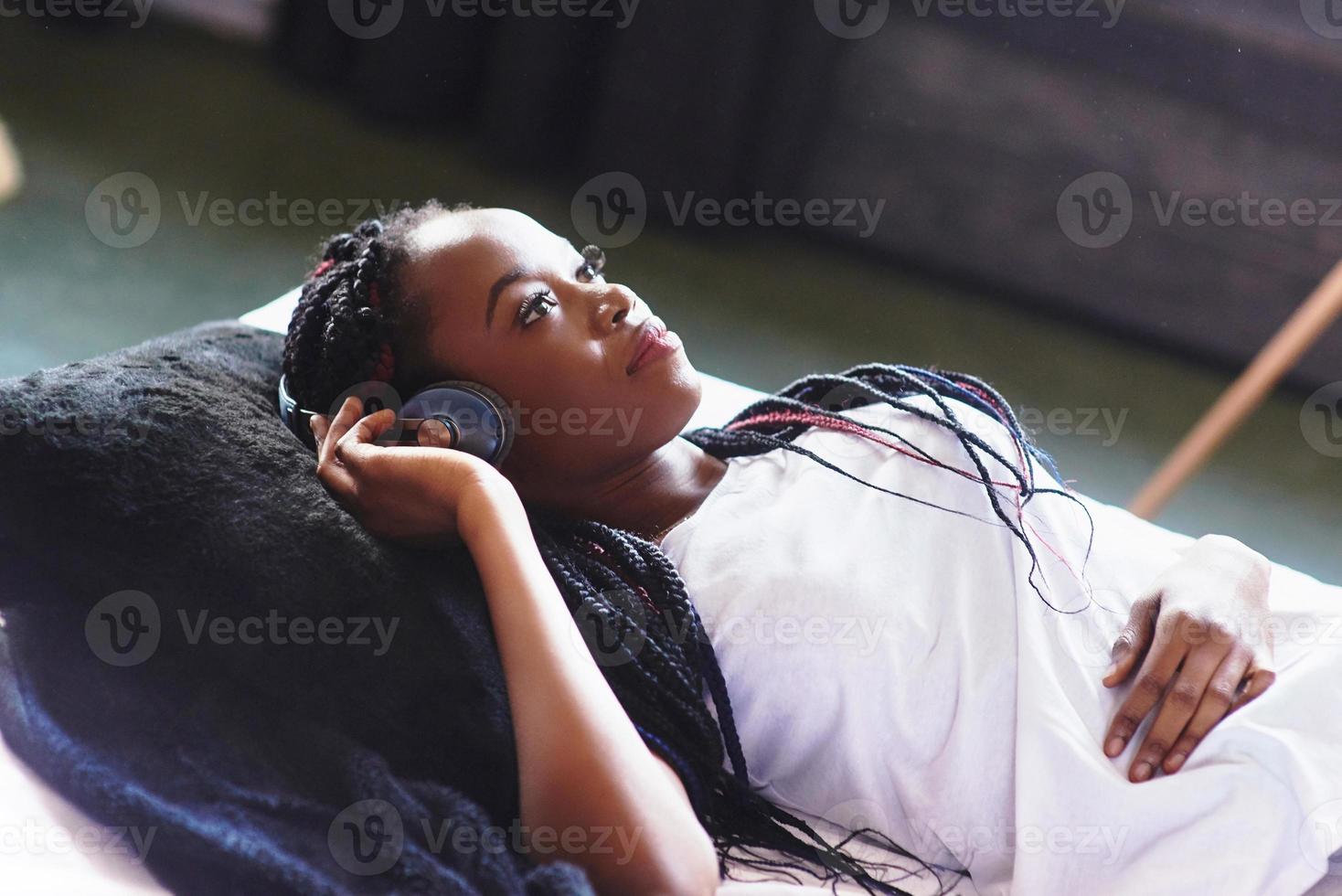 linda garota africana em pijamas sorrindo, ouvindo música em fones de ouvido, deitada na cama. olhos fechados. de cima foto