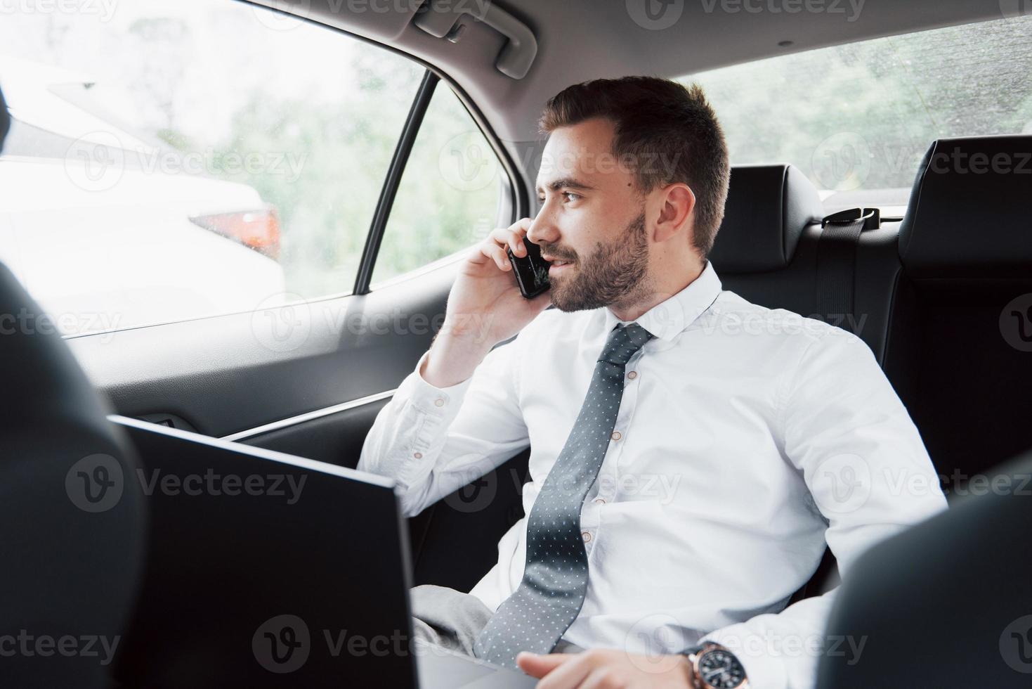 um jovem empresário trabalhando no laptop e falando ao telefone enquanto está sentado na parte de trás do carro. trabalha em movimento, aprecia seu tempo foto
