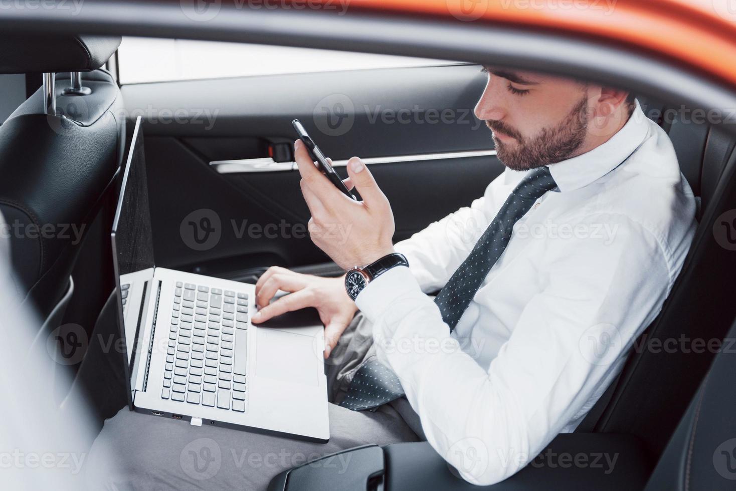 um jovem empresário trabalhando no laptop e falando ao telefone enquanto está sentado na parte de trás do carro. trabalha em movimento, aprecia seu tempo foto