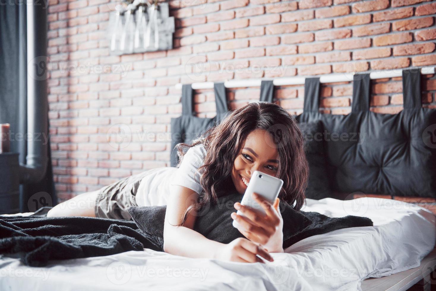 retrato de mulher bonita acordando na cama dela e olha para o telefone. verificar redes sociais, enviar sms. a garota está vestindo uma camiseta foto