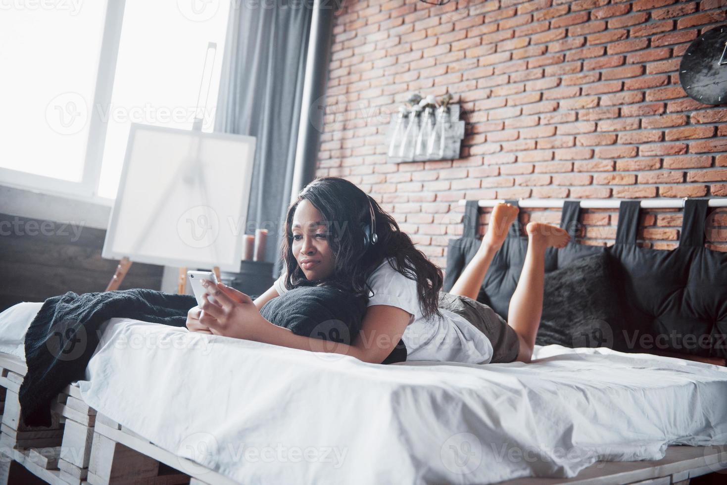 linda garota africana em pijamas sorrindo, ouvindo música em fones de ouvido, deitada na cama foto