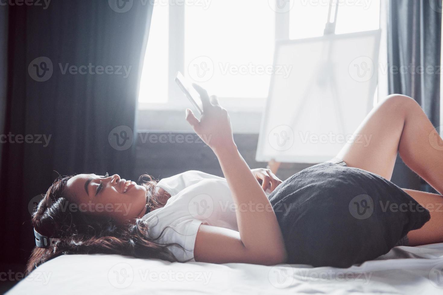 retrato de mulher bonita acordando na cama dela e olha para o telefone. verificar redes sociais, enviar sms. a garota está vestindo uma camiseta foto