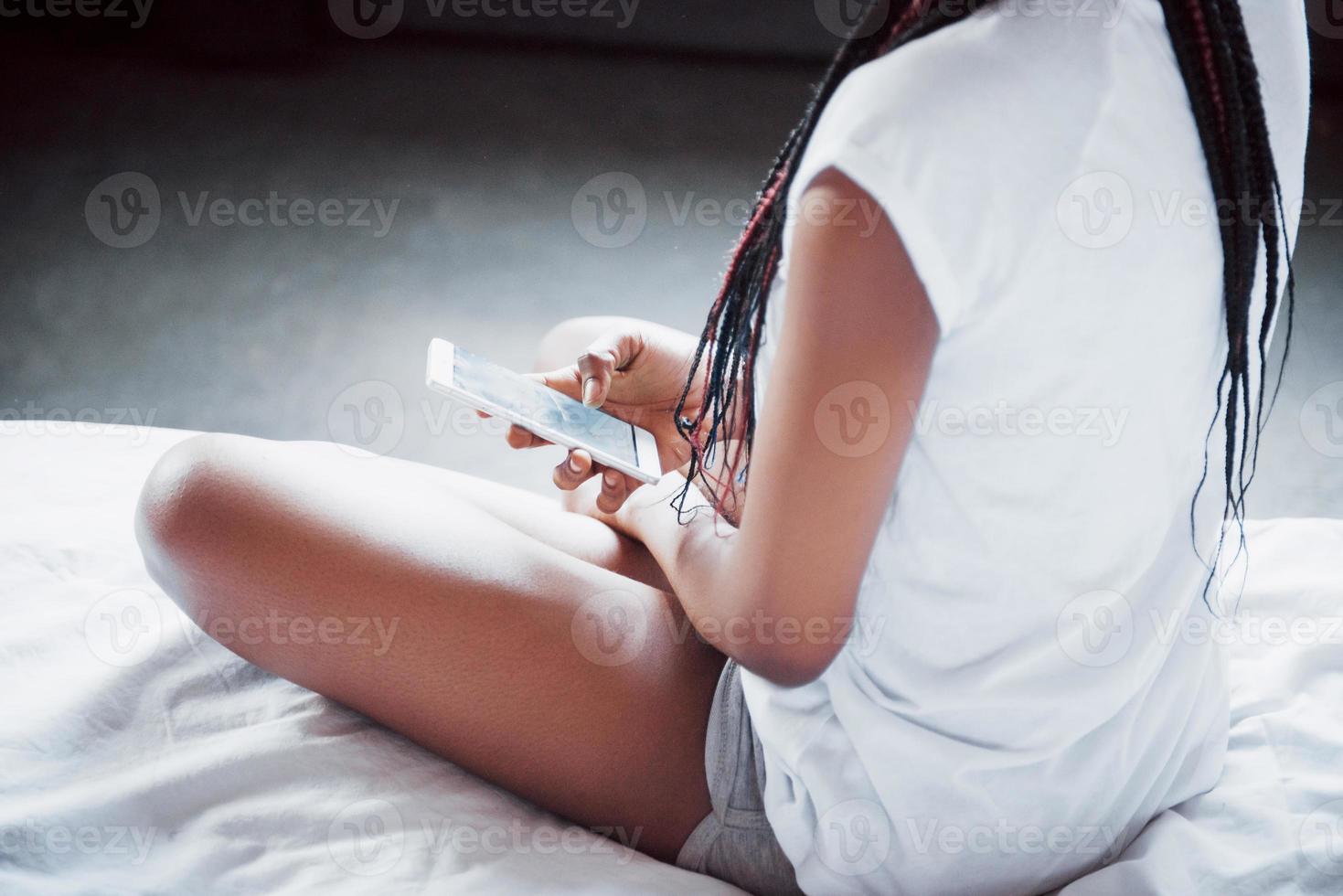 retrato de mulher bonita acordando na cama dela e olha para o telefone. verificar redes sociais, enviar sms. a garota está vestindo uma camiseta foto