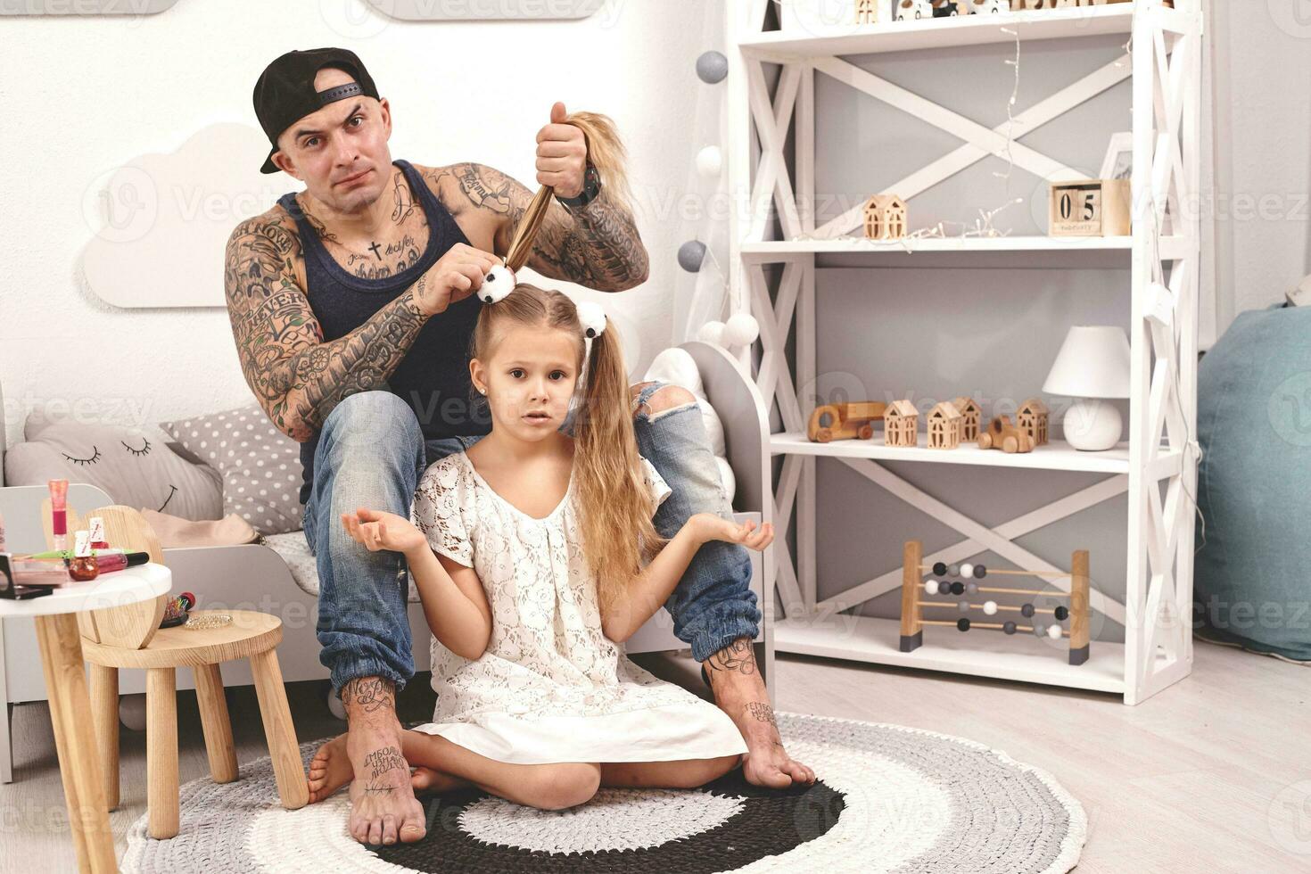 engraçado Tempo tatuado pai dentro uma boné e dele criança estão jogando às lar. Papai é fazendo dele da filha cabelo dentro dela quarto. família feriado e união foto