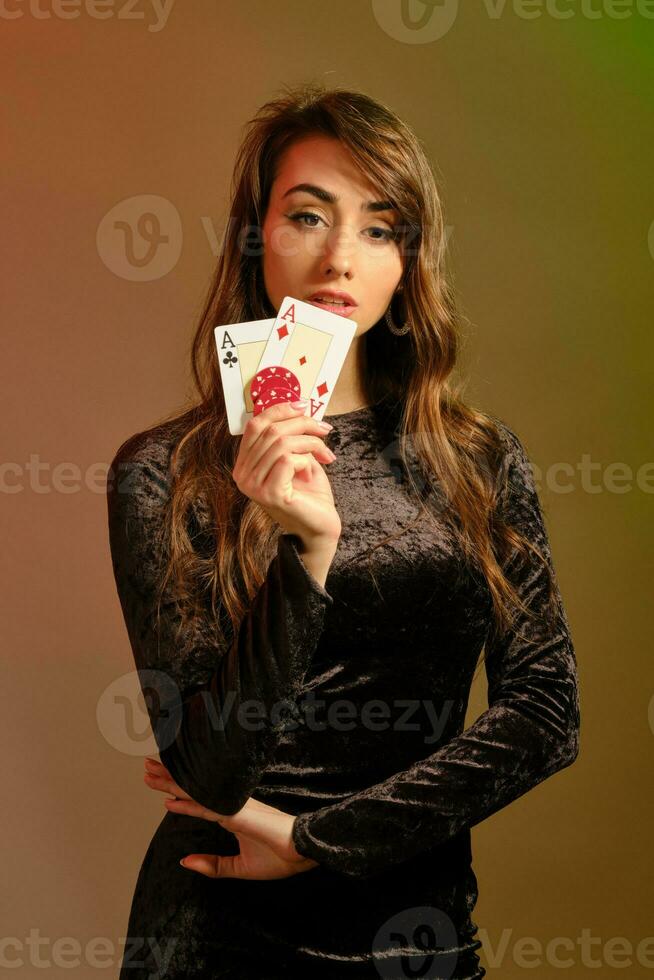 morena fêmea dentro Preto vestir mostrando dois vermelho salgadinhos e ases, posando contra colorida estúdio fundo. jogatina, pôquer, casino. fechar-se. foto