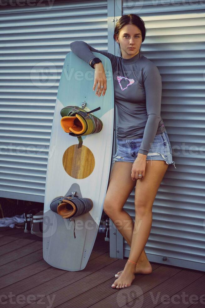 morena menina dentro cinzento gola alta e jeans calção é posando com dela prancha de wakeboard sentado às uma costeiro zona. esporte e lazer. fechar-se. foto