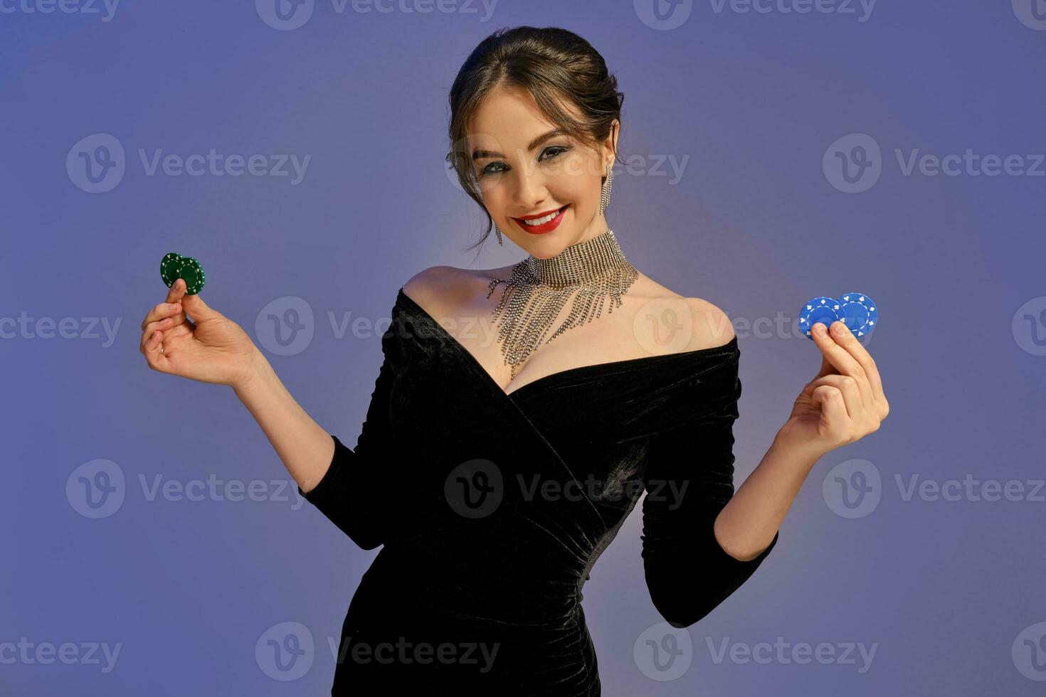 morena mulher dentro Preto vestir e brilhante joia. sorridente, mostrando dois azul e verde salgadinhos, posando em roxa fundo. pôquer, casino. fechar-se foto